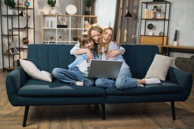 Single Caucasian mother and little children sitting on couch using laptop, family enjoy distant talk by video call, spend time in internet buying, e-commerce users, cartoons or video fun concept
