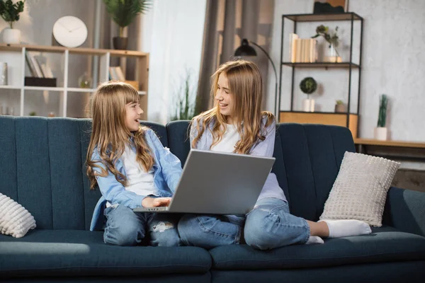 Happy sisters or friends two cute blond girls sitting together on comfortable couch, using laptop, holding video call with father or greeting grandparents, recording video in living room.