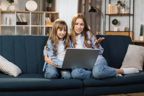 Caucasian cute female children sitting on couch using laptop. Two sisters enjoying distant talk by video call, spend time in internet buying, e-commerce users, watching cartoons or video film.