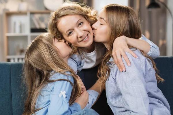 Young little girls child kiss overjoyed middle-aged mom relaxing together on sofa in living room, happy dughters rest enjoy time at home with senior mother, showing love and care.