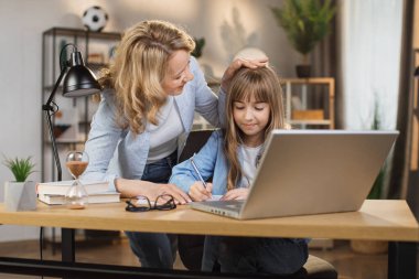 Little daughter learning distance online class on video call virtual lesson with mother, studying at home using laptop computer. Parent or tutor helping child daughter with remote education.