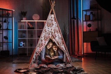 Happy blond woman nanny or older sister reading book to her lovely younger sister inside teepee tent. Comfort at home, evening storytelling and family concept.