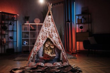 Happy blond woman nanny or older sister reading book to her lovely younger sister inside teepee tent. Comfort at home, evening storytelling and family concept.