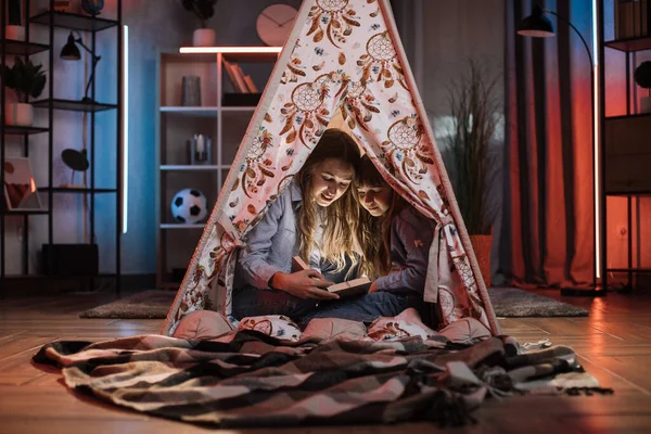 Caring caucasian older sister or nanny with her younger sister inside toy wigwam while reading fairytale. Dark atmosphere at cozy home.