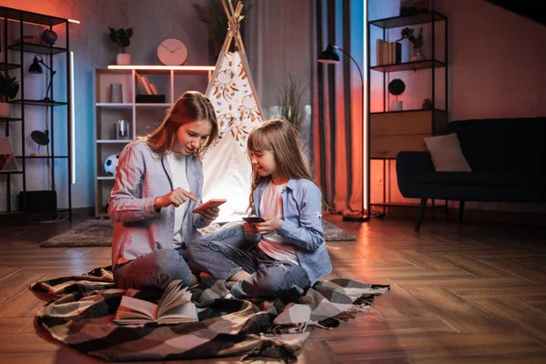 Caring caucasian older sister or nanny with her younger sister outside toy wigwam using their smart phones. Dark atmosphere at cozy home.