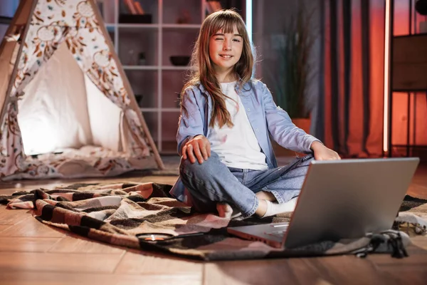Caucasian pretty blond girl sitting on floor in pose of lotos outside wigwam while using laptop for listening relaxing music during evening leisure time. Cozy home concept.