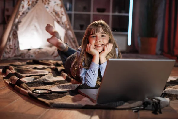 Beautiful cute young girl on floor outside wigwam at home using laptop computer. Happy female spending evening time by watching favorite movie on digital device at home.