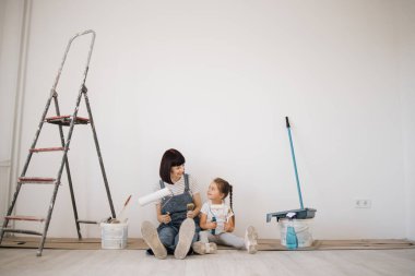 Genç anne ve duvar boyası silindirleri ve fırçalarıyla beyaz duvar arka planında ev tadilatı sırasında yerde oturan tatlı sarışın kız..
