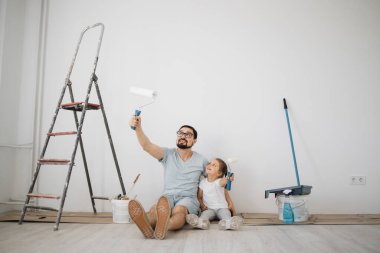 Genç baba ve küçük sarışın kızı duvar boyaları ile yerde oturuyorlar. Evin tadilatı sırasında beyaz arka planda..