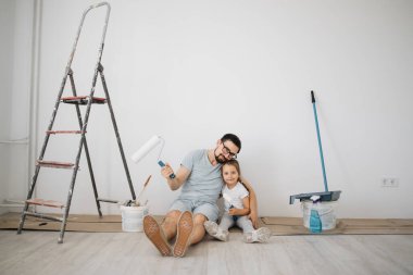 Neşeli beyaz adam babası ve tatlı küçük kızı ellerinde boya rulolarıyla beyaz ışık odasında yerde oturuyorlar..