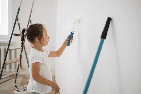 Repair in the apartment. Happy cute child girl painting the wall with paint roller standing in a bright room. Home repairs concept.