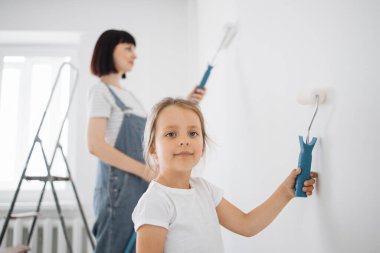 Gülümseyen 4 yaşındaki bir kız elinde boya silindiri tutuyor. Çocuk, arkasında duran annesine yardım ediyor, duvarın yanında, onarımda..