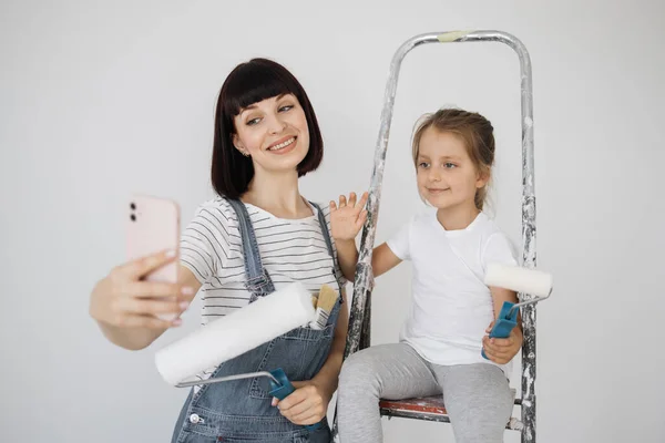 A cheerful happy family is renovating a purchased apartment, mom holding smart phone. The mother hugs her daughter, sitting on a ladder and having video call.