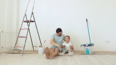 Genç baba ve küçük sarışın kızı duvar boyaları ile yerde oturuyorlar. Evin tadilatı sırasında beyaz arka planda..