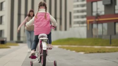 Anaokulunun arka planında sonbahar kıyafetleri giyen sevimli bir kızın şehir binalarının arka planında genç ve güzel annesine bisiklet sürüşünün görüntüsü..