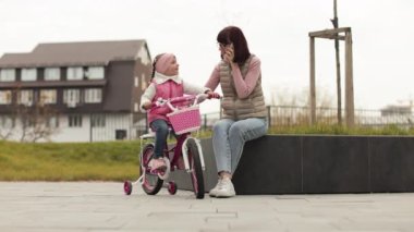 Mutlu aile tatilleri, genç anne şehir arka planında bankta oturuyor ve anaokulundaki küçük kızı bisiklet sürmeyi öğretirken telefonda konuşuyor..
