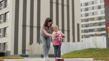 Güzel ve mutlu genç bir anne, küçük tatlı kızına şehir binalarının arka planında, evinin yakınında scooter sürmeyi öğretiyor..