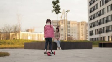 Anaokulunun arka planında, sonbahar kıyafetleri giyen hoş bir kızın, şehir binalarının arka planında oturan genç ve güzel annesine scooter sürüşünün görüntüsü..