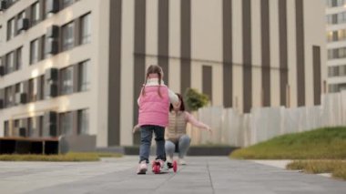 Sonbahar sıcağı içindeki küçük şirin anaokulu kızının kentsel arka plandaki güzel annesinin evine scooter sürüşünü yakından izleyin..