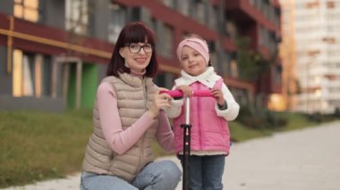 Güzel ve mutlu bir annenin, kızına scooter sürmeyi öğretirken çekilmiş portresi. Çekici beyaz kadın küçük kızına sarılıyor, scooter 'da oturuyor, el sallıyor ve kameraya bakıyor.