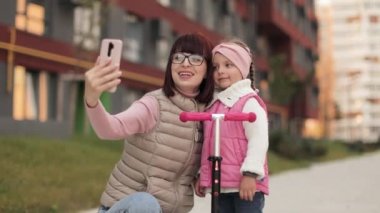 Çekici ve mutlu genç bir anne şirin kızına scooter sürmeyi öğretiyor, gülümsüyor ve cep telefonuna bakıyor, video görüşmesi yapıyor ya da kentin arka planında selfie çekiyor..