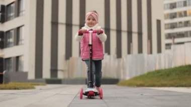 Sonbahar sıcağı içindeki küçük şirin anaokulu kızının kentsel arka planda evinin yanında scooter sürüşünü yakından izle..