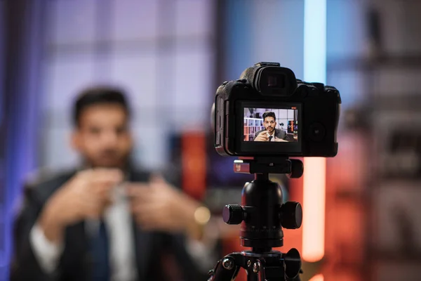 View from camera screen of confident indian bearded businessman, broker in suit sitting in front of camera in evening office during recording video for business vlog.