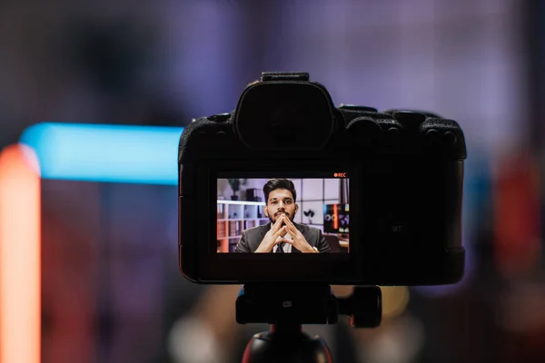 View from camera screen of confident indian bearded businessman, broker in suit sitting in front of camera in evening office during recording video for business vlog.