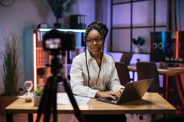 Confident african american businesswoman, broker in white shirt sitting in front of camera in evening office during recording video for business vlog.