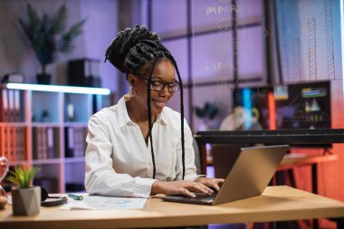 Kendine güvenen Afro-Amerikan finansal uzman ofis çalışanının resmi olarak masada oturan portresi akşam modern ofis laptopuyla çalışıyor..