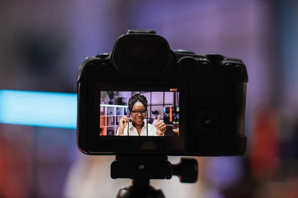 Focus on camera screen with attractive confident experienced smart female african american office manager in headset recording video vlog for internet audience in evening office showing thumb up.