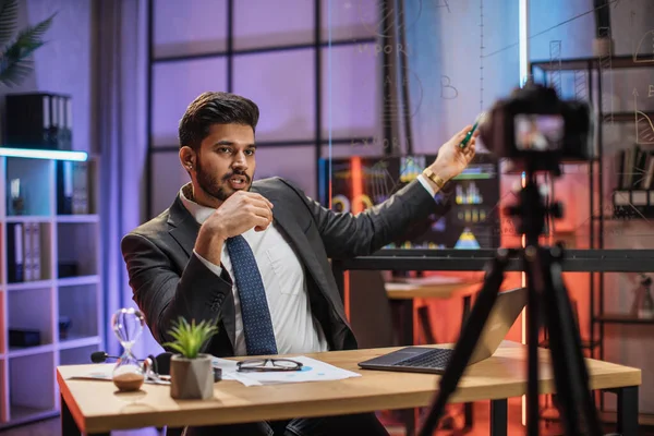 Concept of distant work at night. Attractive confident arab skilled bearded businessman explaining online economic charts on glass board to his colleagues sitting in front of video camera.