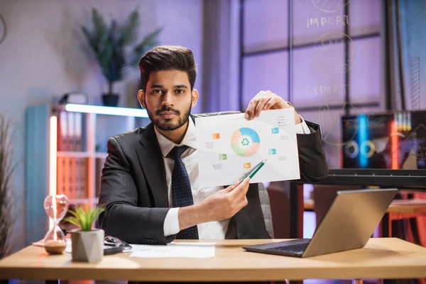 Confident arab financial expert office worker in suit working on laptop, showing paper report with graphs and charts during video meeting with colleagues in office.