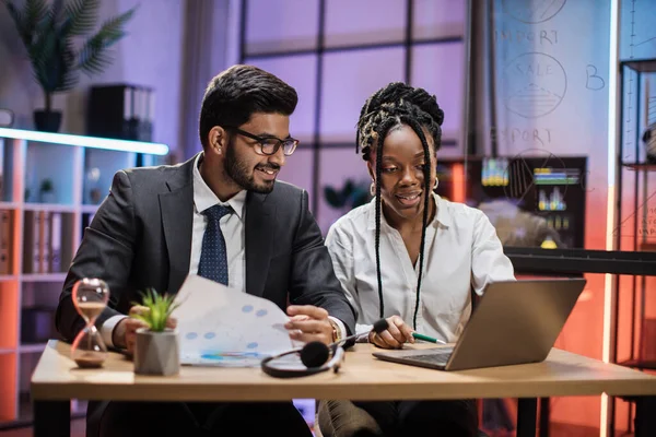 Two multicultural company workers arab financial expert and african woman office worker using laptop, showing paper report with graphs and charts during video meeting with colleagues in office.