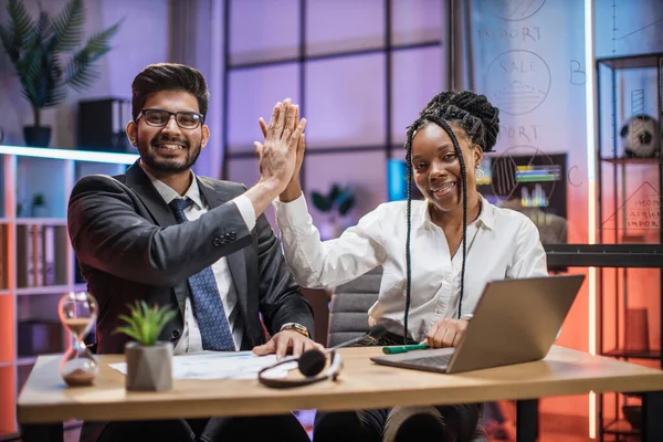 Two multi ethnic economists giving high five, young smiling experienced african american female and arab male office manager in formal wear sitting at table using laptop in evening office.