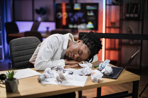 Portrait of tired overworked african american businesswoman in formal wear, sleeping on desk with sheets of crumpled papers in evening office.