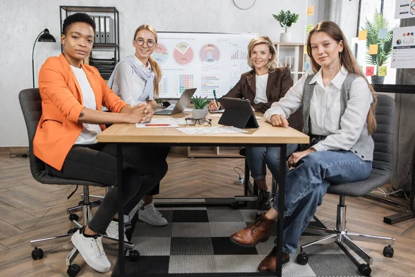 Female economists in formal wear looking at camera while sitting together at office desk with modern gadgets and documents. Multi ethnic women brokers spending evening for examining trade market.