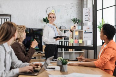 Bir grup yetenekli kadın ekonomist ofis odasında toplanırken iş fikirlerini tartışıyor. Cazibeli genç bir kadın, çeşitli grafik ve çizelgelerle cam tahtanın yanında duruyor..