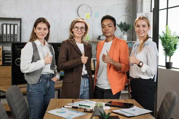 Portrait of female economists in formal wear looking at camera while standing together at office with graphs and charts. Multi ethnic brokers spending time for examining trade market showing thumb up