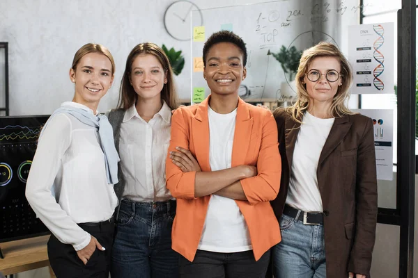 Close up portrait of female economists in formal wear looking at camera while standing together at office with graphs and charts. Multi ethnic brokers spending time for examining trade market.