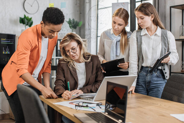 Teamwork, brainstorming in office. Diverse multiracial business people working together with laptop and tablet pc while their senior woman leader sign in report papers.