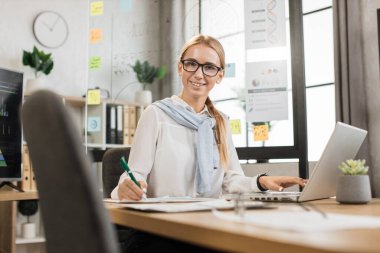 Şık bir şekilde giyinmiş çekici bir kadının portresi ofis masasında otururken notlar alır ve modern dizüstü bilgisayar kullanır. Güzel bir şirket çalışanı, sarı saçlı, kameraya bakıyor..