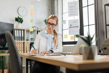 Beyaz gömlekli çekici bir kadının portresi notlar yazıyor ve ofis masasında otururken modern laptop kullanıyor. Güzel bir şirket çalışanı. Sarı saçlı. Proje raporlarına bakıyor..