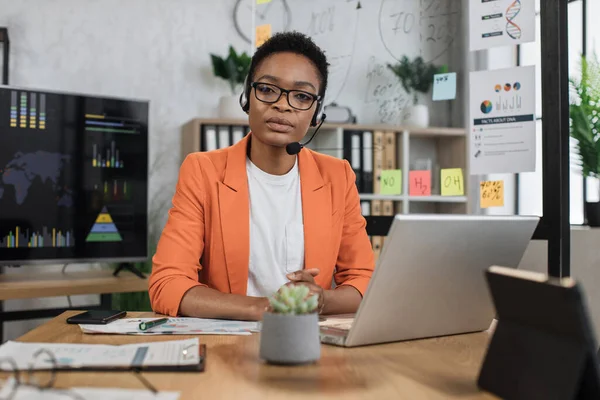Portrait of attractive african business woman in headset sitting at office desk and looking at camera. Business lady having video conference. Business meeting online.
