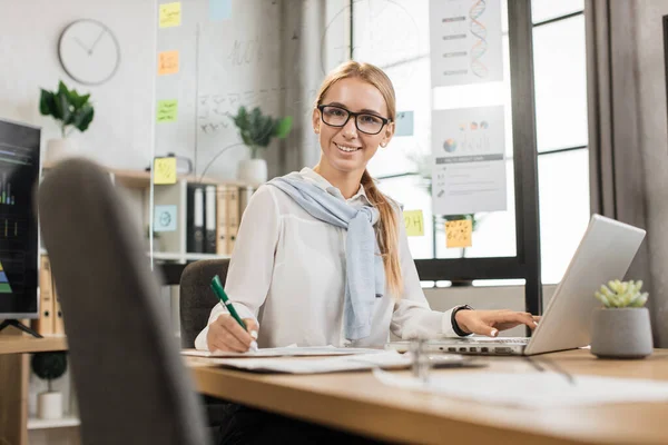 Portrait of charming woman in stylish formal wear writing notes and using modern laptop while sitting at office desk. Pretty company worker with blond hair looking at camera.