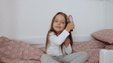 5 yaşında bir kız sabah yatak odasında uzun saçlarını tarıyor. Saçını tarayan komik bir kız çocuğu. Mutlu bebek yatakta oturuyor..