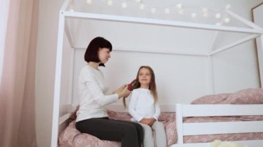 Gülümseyen genç bir bayan anaokulu kızının uzun, pürüzsüz saçlarını tarıyor, yatak odasının içinde yatakta oturuyor. Anne, kız ve güzellik hizmetleri, aile ilişkileri..