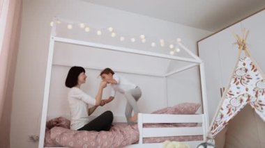 Neşeli anne bebek bakıcısı şirin, aktif, küçük bir kızla oynuyor. Yatağa atlıyor, kaygısız anne ve küçük kız el ele tutuşuyor ve gülüyor. Modern yatak odasında evde eğleniyor.