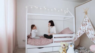 Güzel, genç bir anne ve pijamalı küçük kızının evde yatakta birbirlerine hava öpücüğü verirken çekilmiş uzun bir portresi. Sevimli kız ve annesi aile gününün tadını çıkarıyorlar..