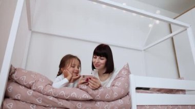 Güzel genç kadın ve büyüleyici küçük kızı kulaklık bir akıllı telefon kullanarak ve evde yatakta yatan süre gülümseyen müzik dinleme